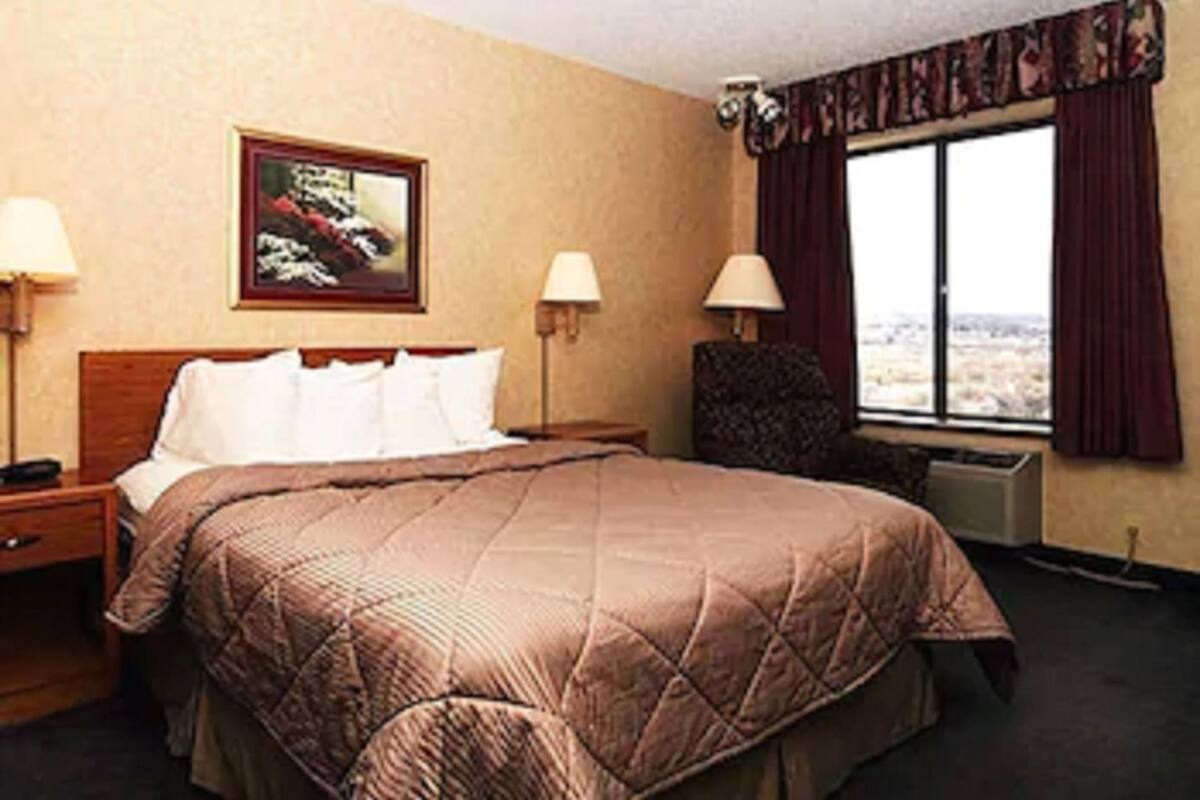 Una habitación de hotel con una cama grande, dos mesitas de noche, una silla, una ventana con cortinas y un cuadro en la pared.