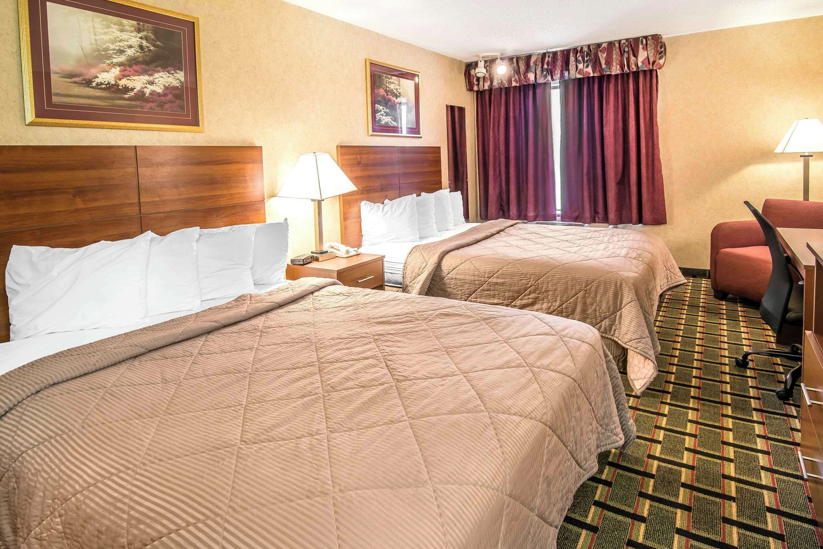 A hotel room with two beds, a wooden headboard, a desk with a chair, a telephone, and a window with curtains.