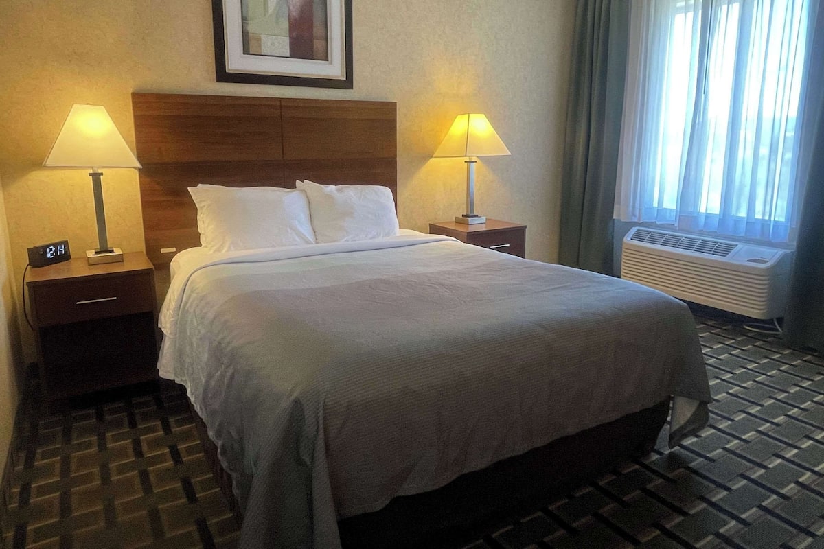 Una habitación de hotel con una cama grande, dos mesitas de noche con lámparas, un reloj y una ventana con cortinas.