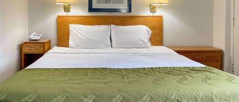 Una cama bien hecha con una colcha verde, dos lámparas de noche, un cabecero de madera, una impresión botánica enmarcada en la pared y un teléfono en la mesita de noche.