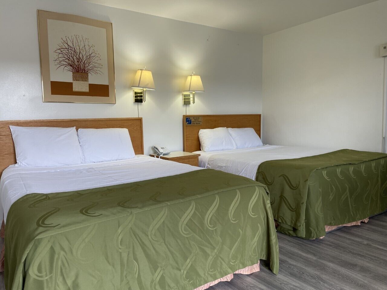 Ein Hotelzimmer mit zwei Betten, jeweils mit grüner Bettdecke, einem hölzernen Kopfteil und einem Nachttisch mit Lampe.
