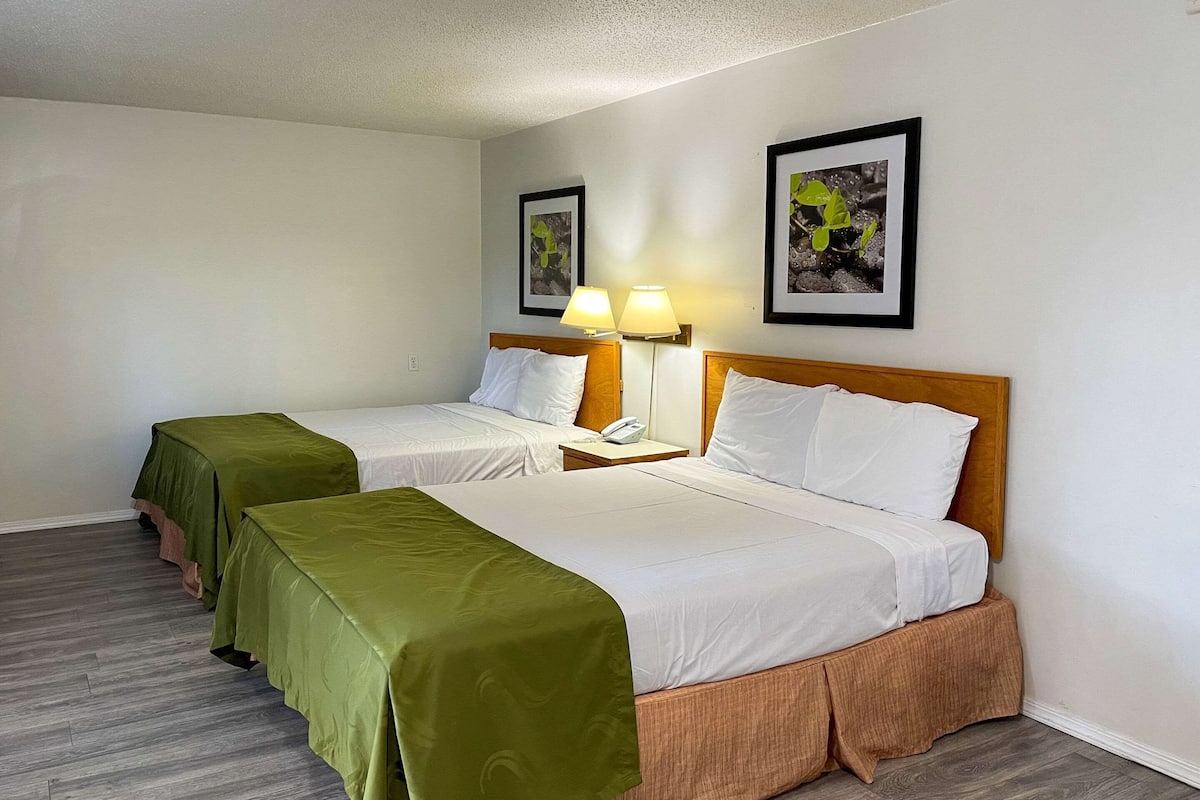 A hotel room with two beds, each with a green bedspread, white linens, and wooden headboards. There are two bedside tables with lamps, and artwork is displayed on the walls.
