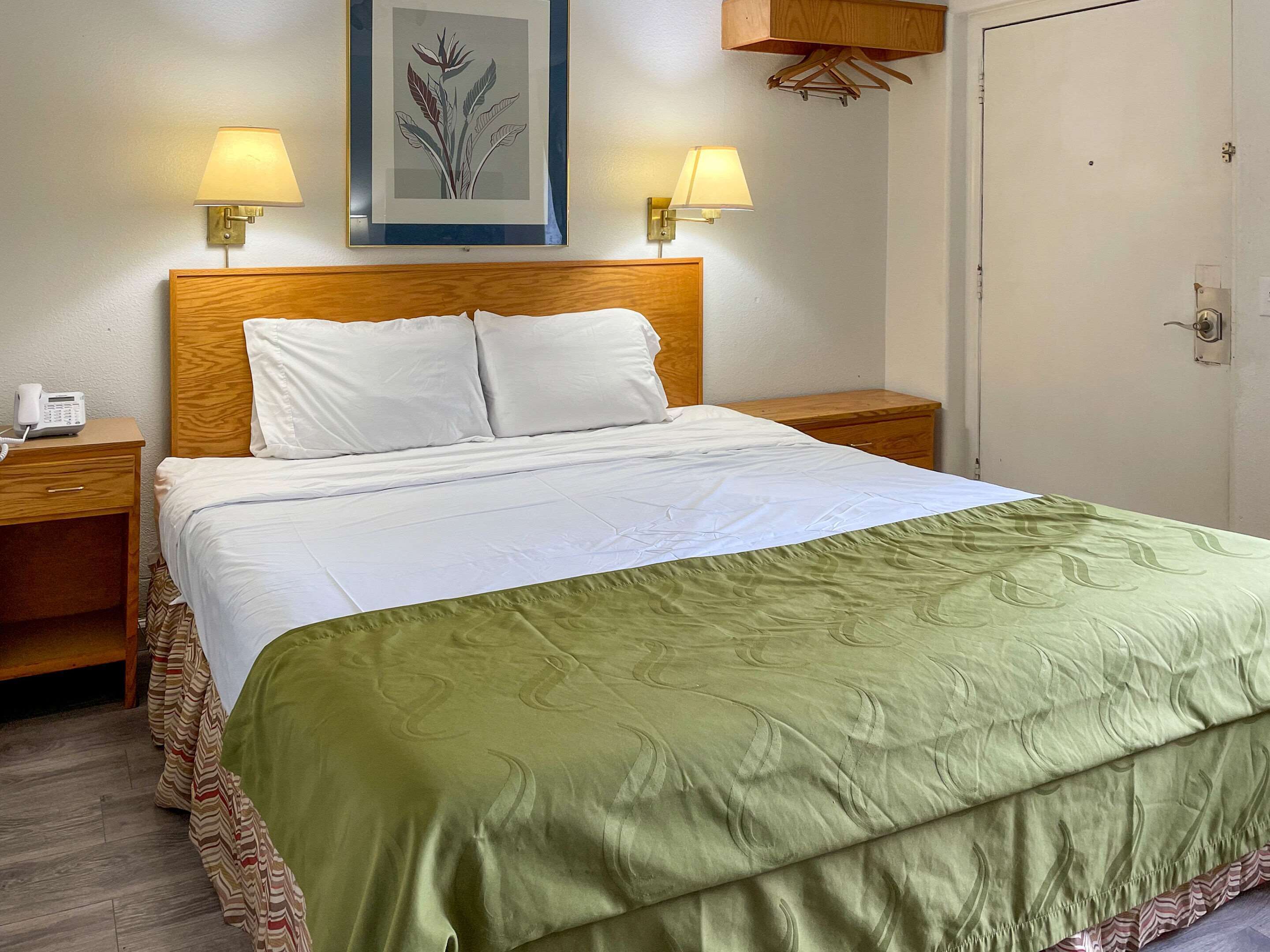 Una cama bien hecha con una colcha verde, dos lámparas de noche, una obra enmarcada sobre la cama y un cabecero de madera.