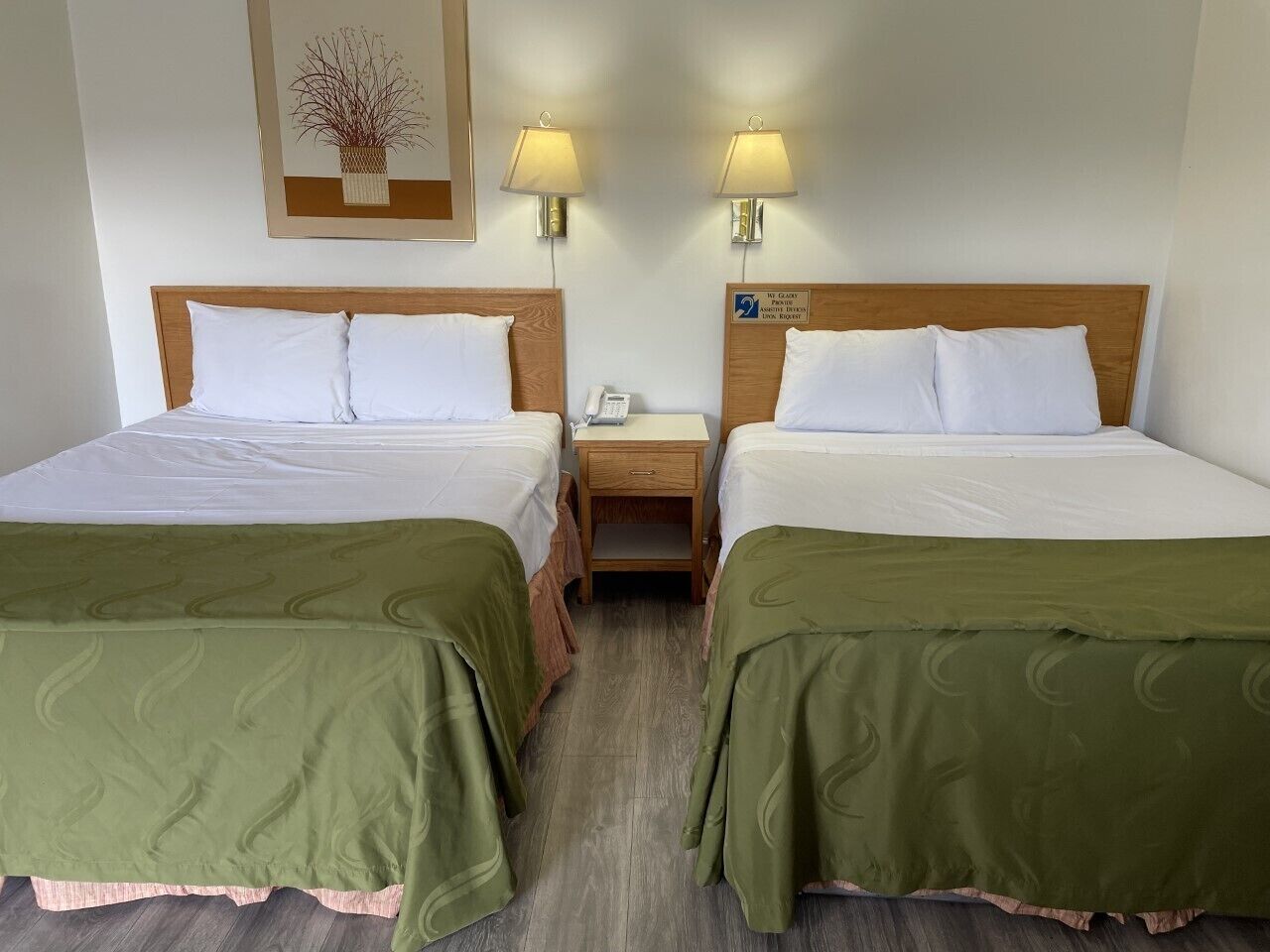 A double room with two beds, each with a wooden headboard and green bedspreads. There are bedside tables with lamps and a small nightstand with a phone.