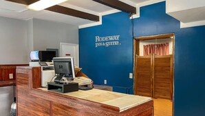 Un mostrador de recepción con una computadora, una caja registradora y un teléfono. En la pared hay un letrero que dice "RODEWAY INN & SUITES".