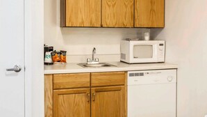 Una cocina compacta con armarios de madera, una encimera blanca, microondas, fregadero y lavavajillas.