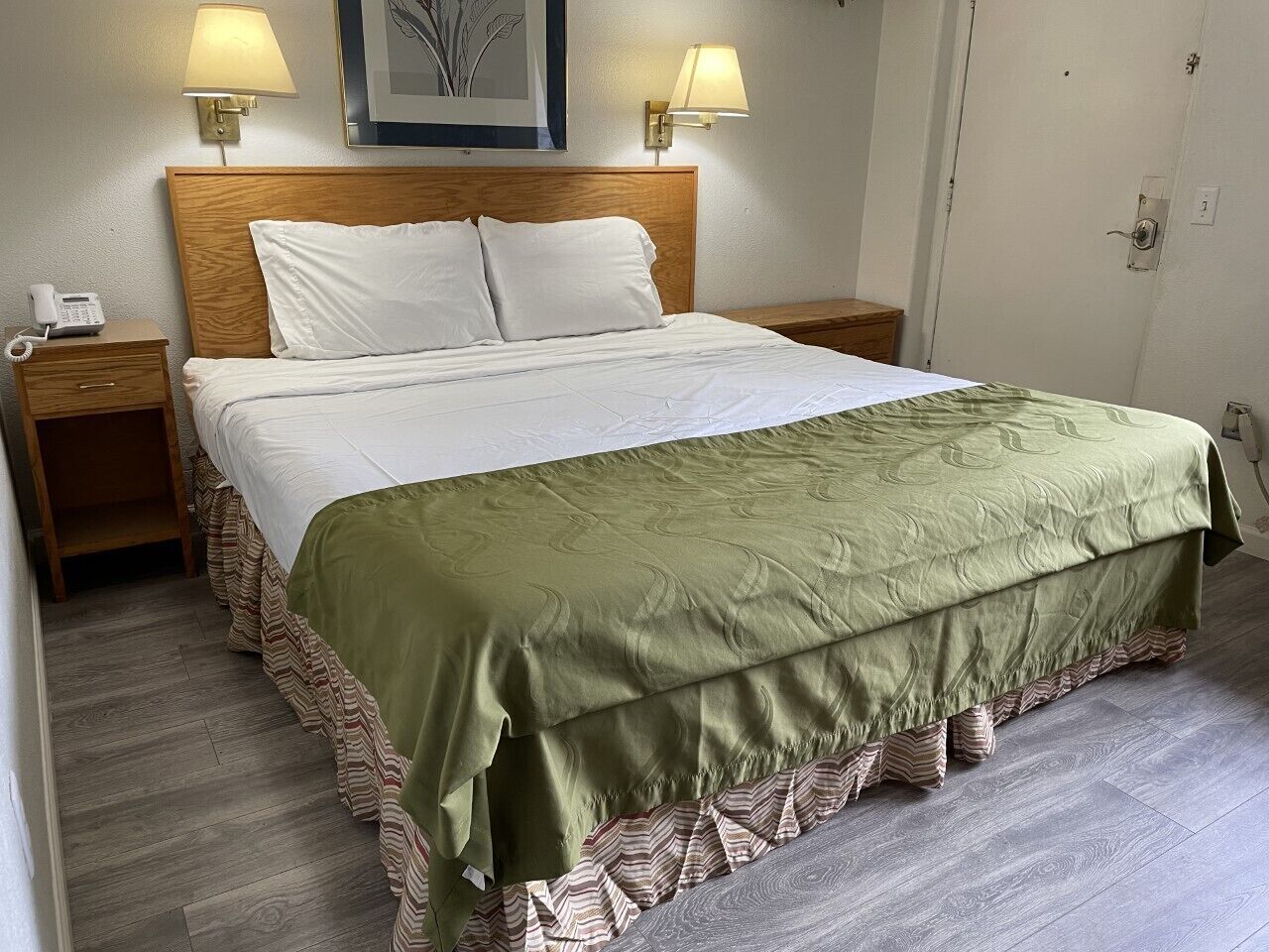 Una cama bien hecha con una colcha verde y almohadas blancas, dos lámparas de noche, una mesita de noche de madera y un cuadro en la pared.