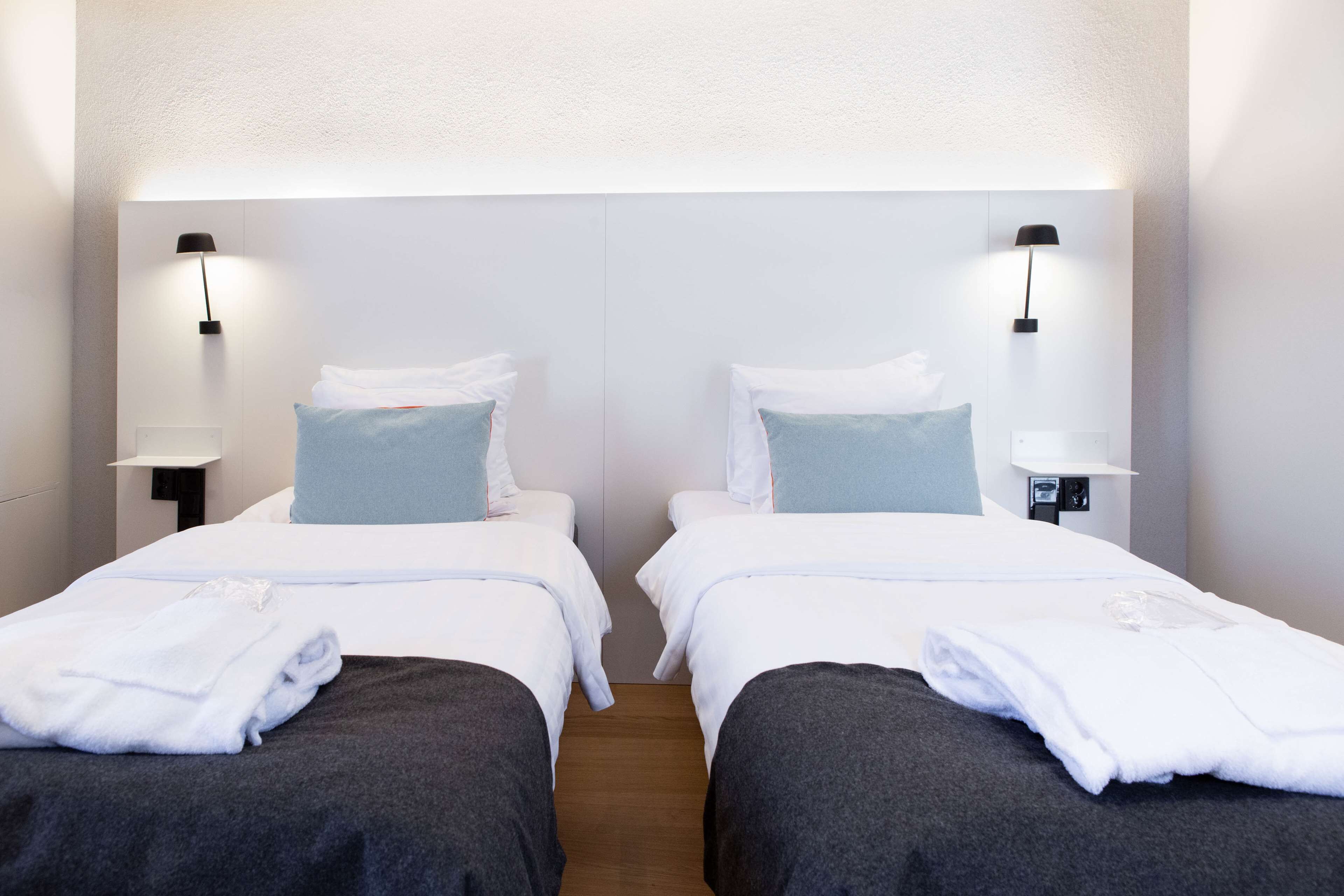 Quarto de hotel com duas camas, cada uma com roupa de cama branca e almofada azul. Há duas mesas de cabeceira com luminárias.