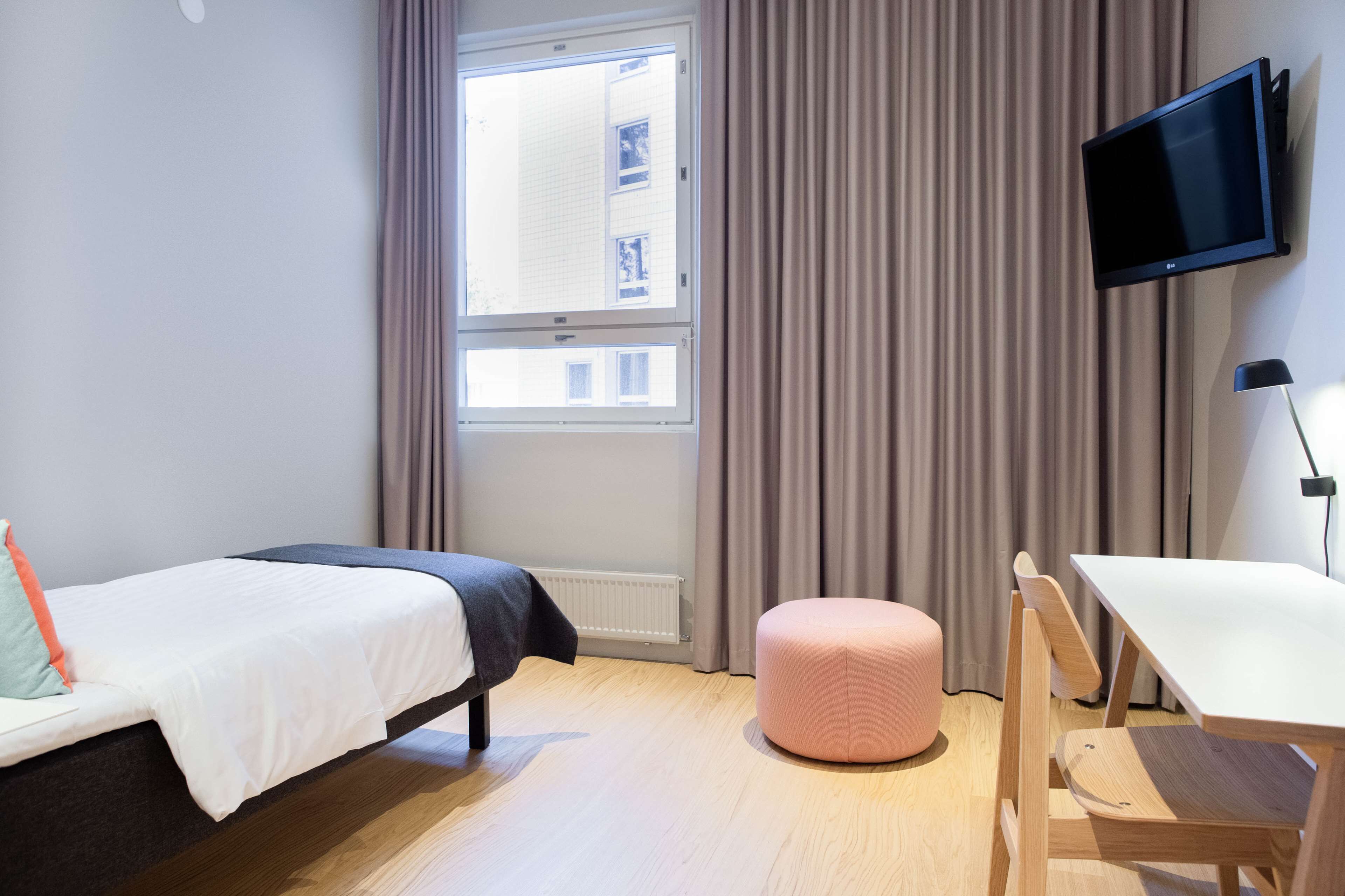 Habitación de hotel con cama, escritorio, un reposapiés rosa, televisor montado en la pared y ventana con cortinas.