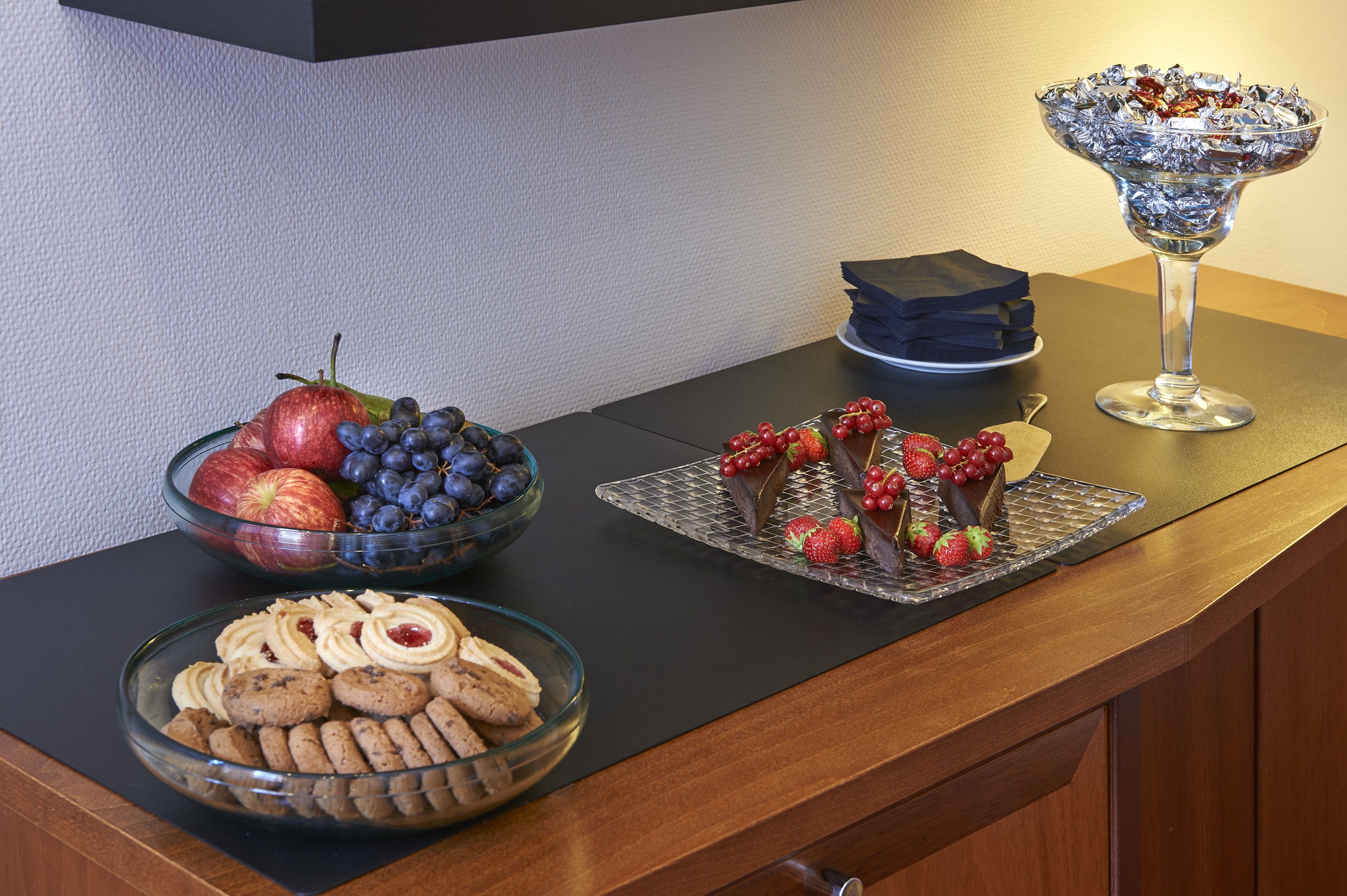 Una mesa con diversos alimentos, incluyendo un tazón de manzanas y uvas, una bandeja de galletas y un recipiente de vidrio con chocolate y fresas.