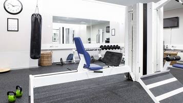 Uma academia com um saco de pancadas, uma cadeira de exercícios azul, halteres e kettlebells, e um espelho.