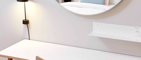A hotel room with a white desk, a wooden chair, a bedside table, a lamp, and a mirror reflecting a bed with a pillow.