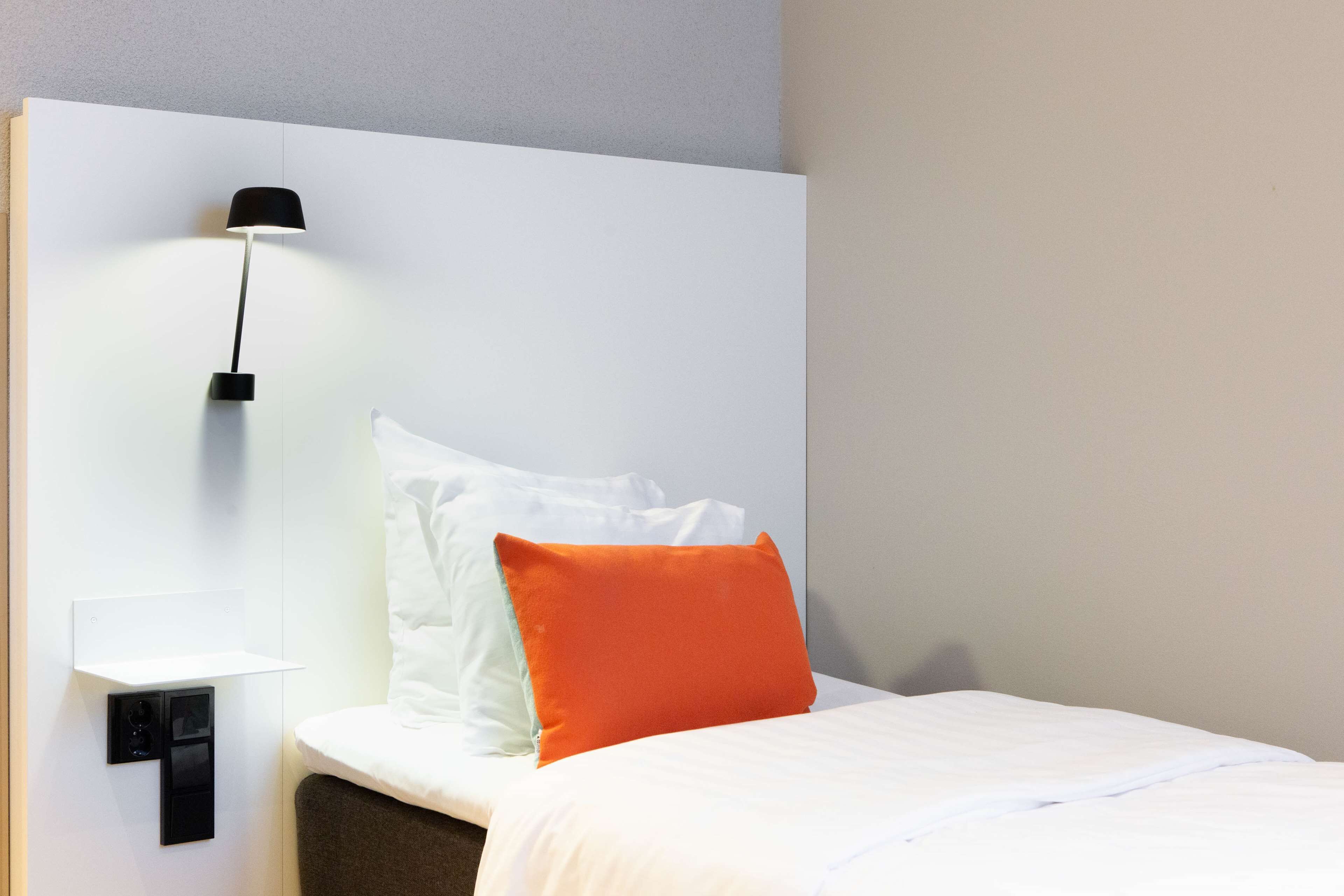 Una cama bien hecha con almohadas blancas y anaranjadas, una lámpara negra fijada a la pared y una mesita de noche blanca.