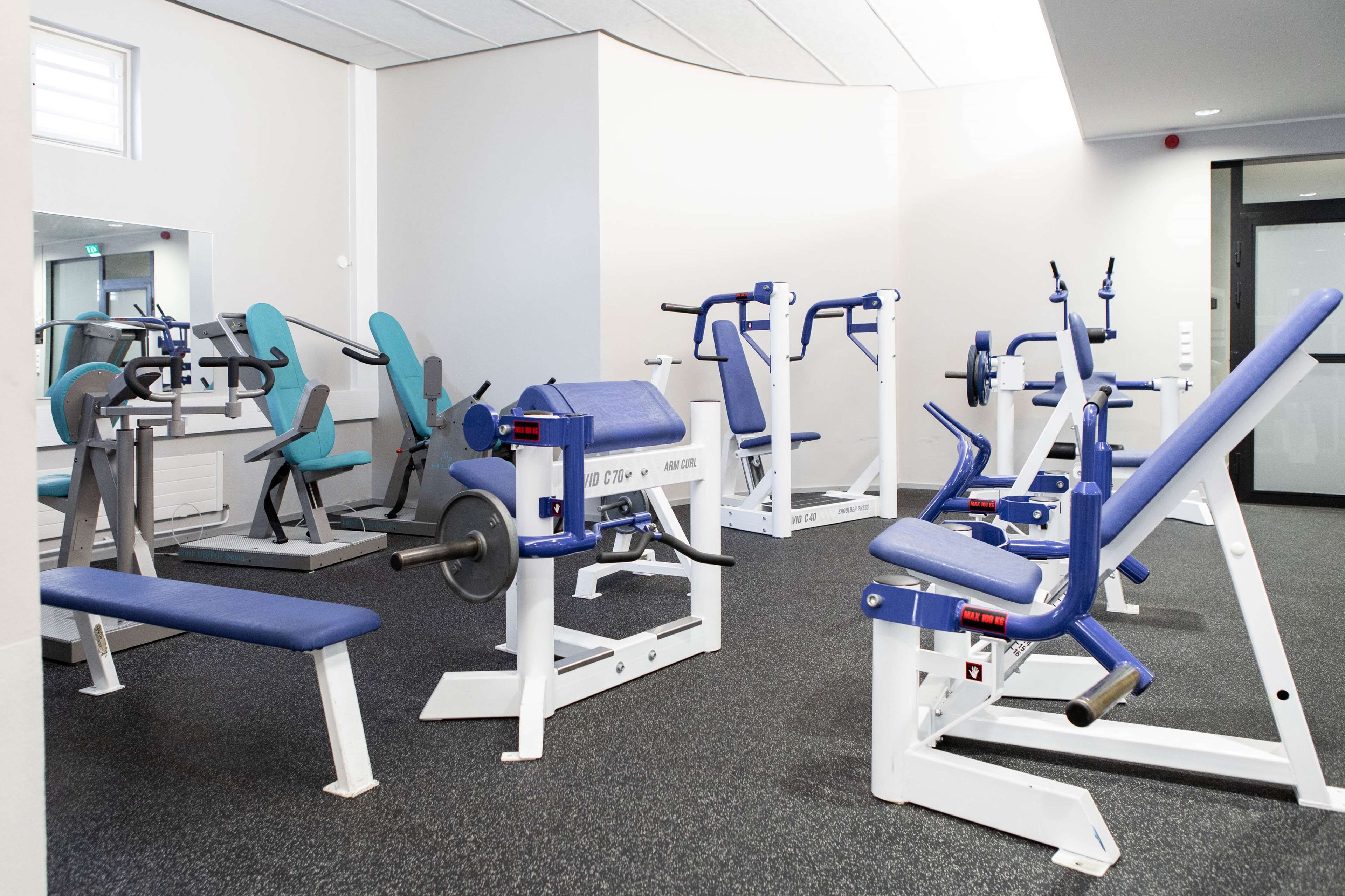 Un gimnasio con diferentes máquinas de ejercicio, una banca y una puerta al fondo.