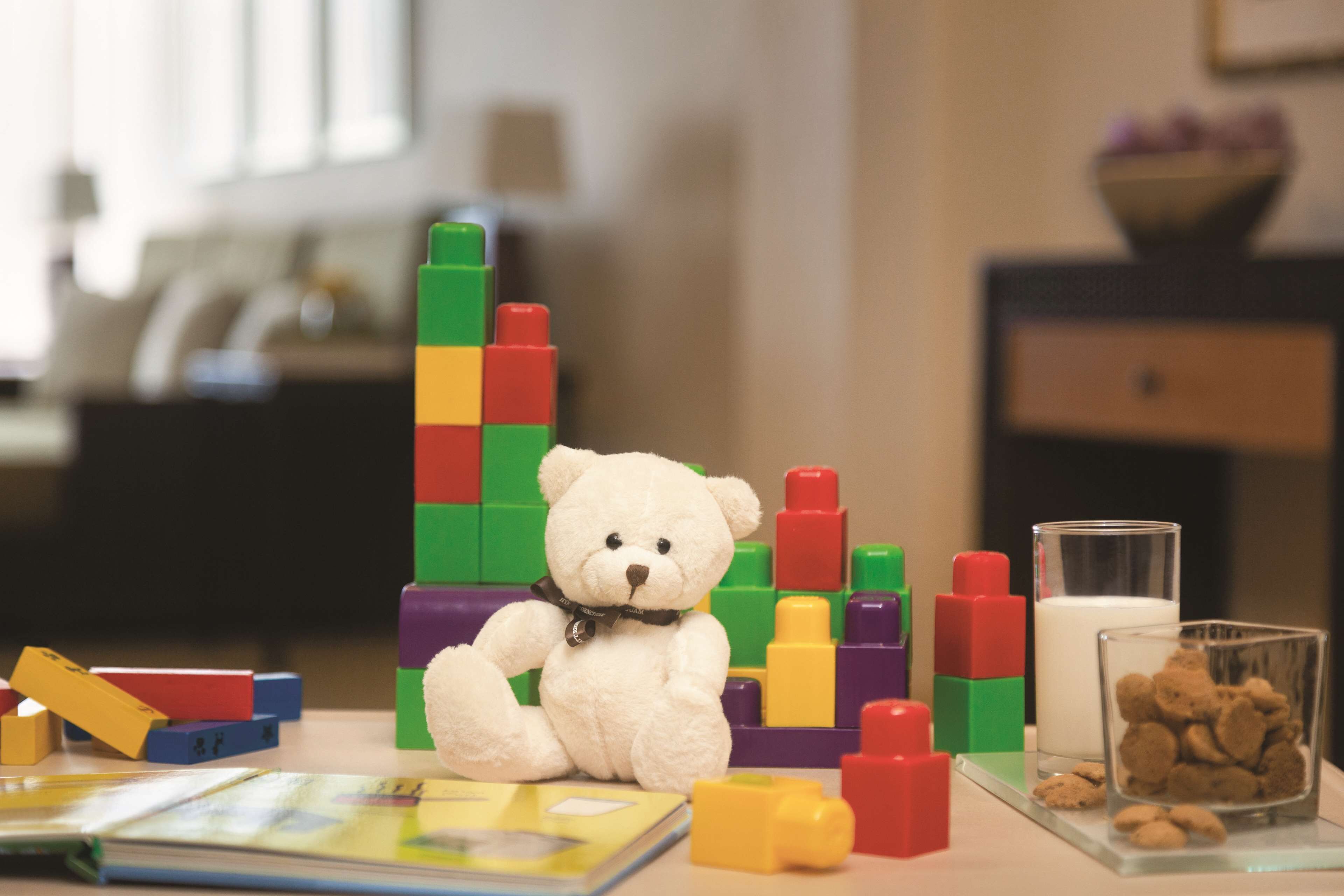A white teddy bear sitting among colorful building blocks, a glass of milk, and a bowl of cereal on a table.