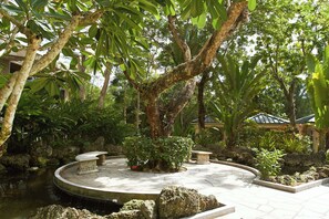 A circular stone patio with a central fountain, surrounded by lush greenery and a small pond.