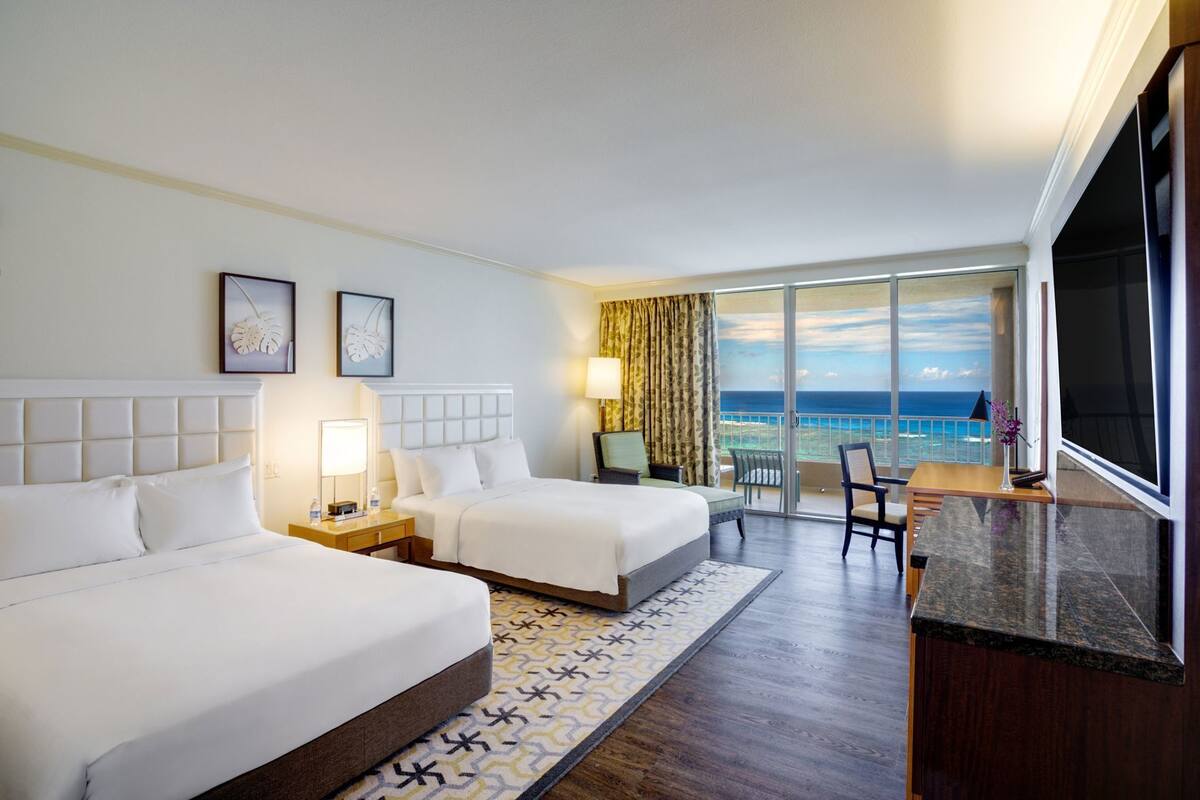A hotel room with two beds, a desk, a chair, a TV, and a balcony with a view of the ocean.