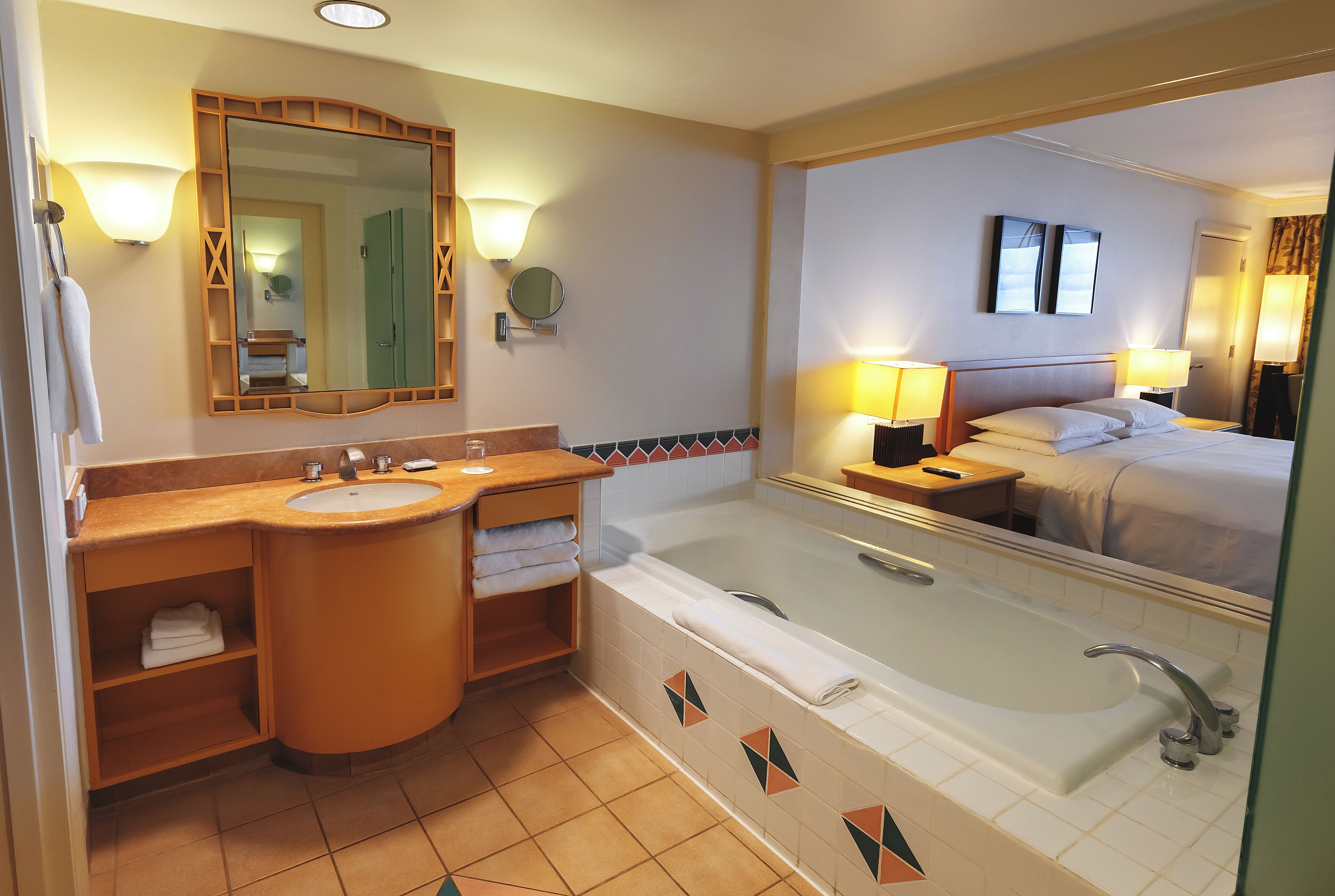 A hotel bathroom with a large bathtub, two sinks, a mirror, and a nightstand with a lamp.
