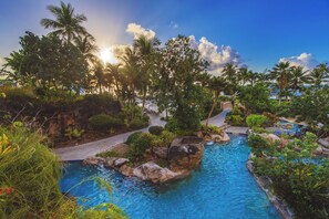 Un área de piscina con vegetación tropical y palmeras, rodeada de rocas y un cielo despejado.