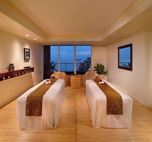 A room with two beds, a large window with a view, a small table, and a shelf with items.