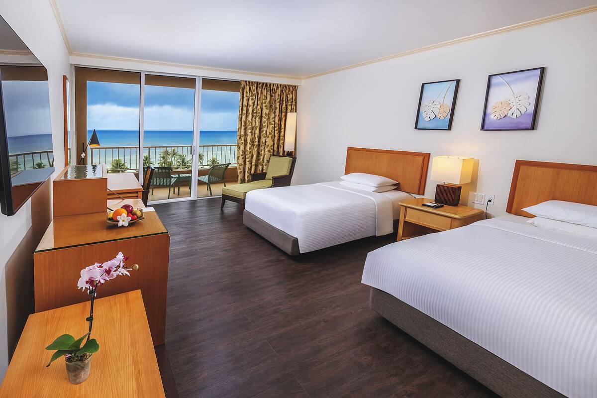 A hotel room with two beds, a desk, a chair, a TV, and a balcony with a view of the ocean.