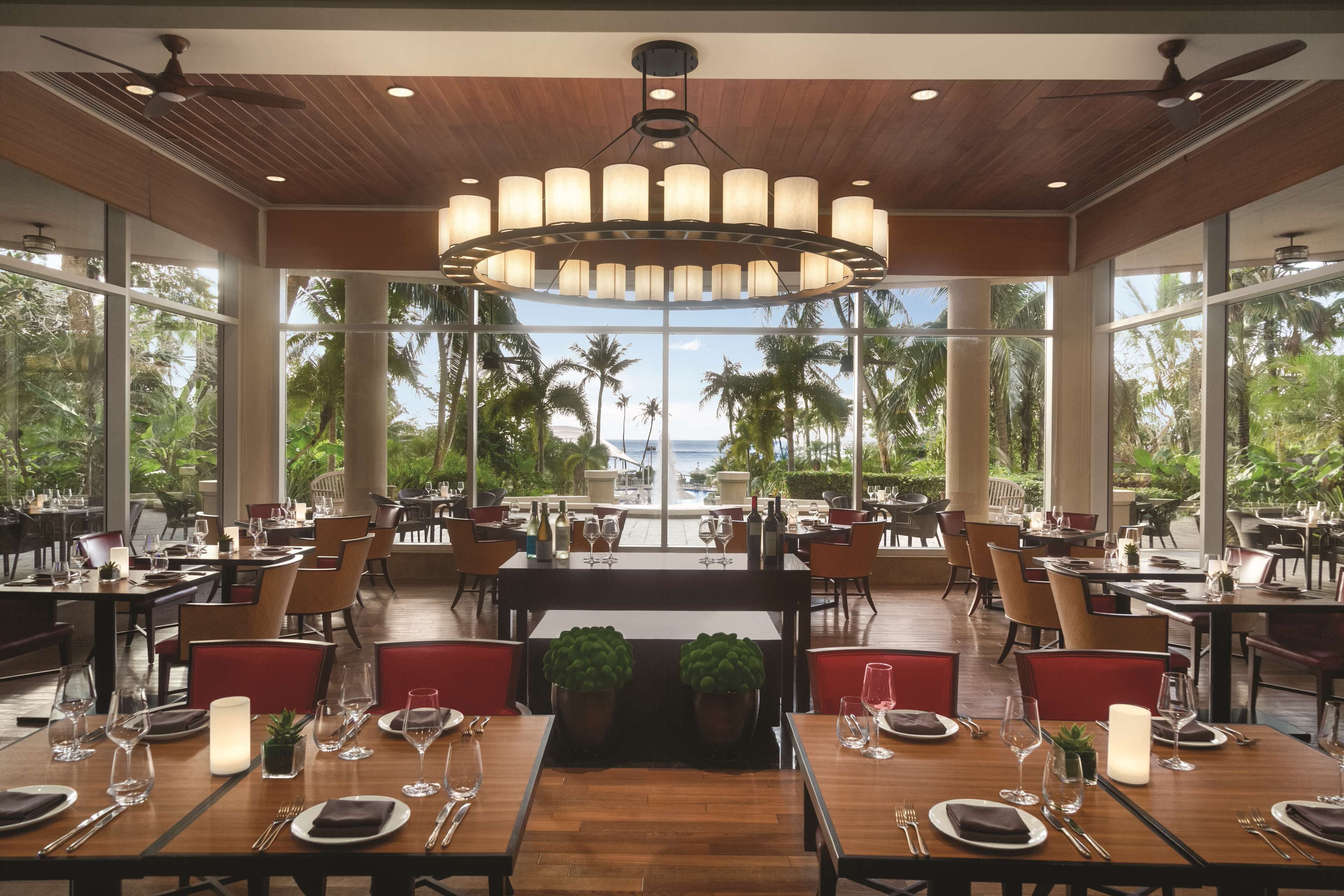 A restaurant with wooden tables, red chairs, and a large chandelier. It has large windows with a view of palm trees and the ocean.
