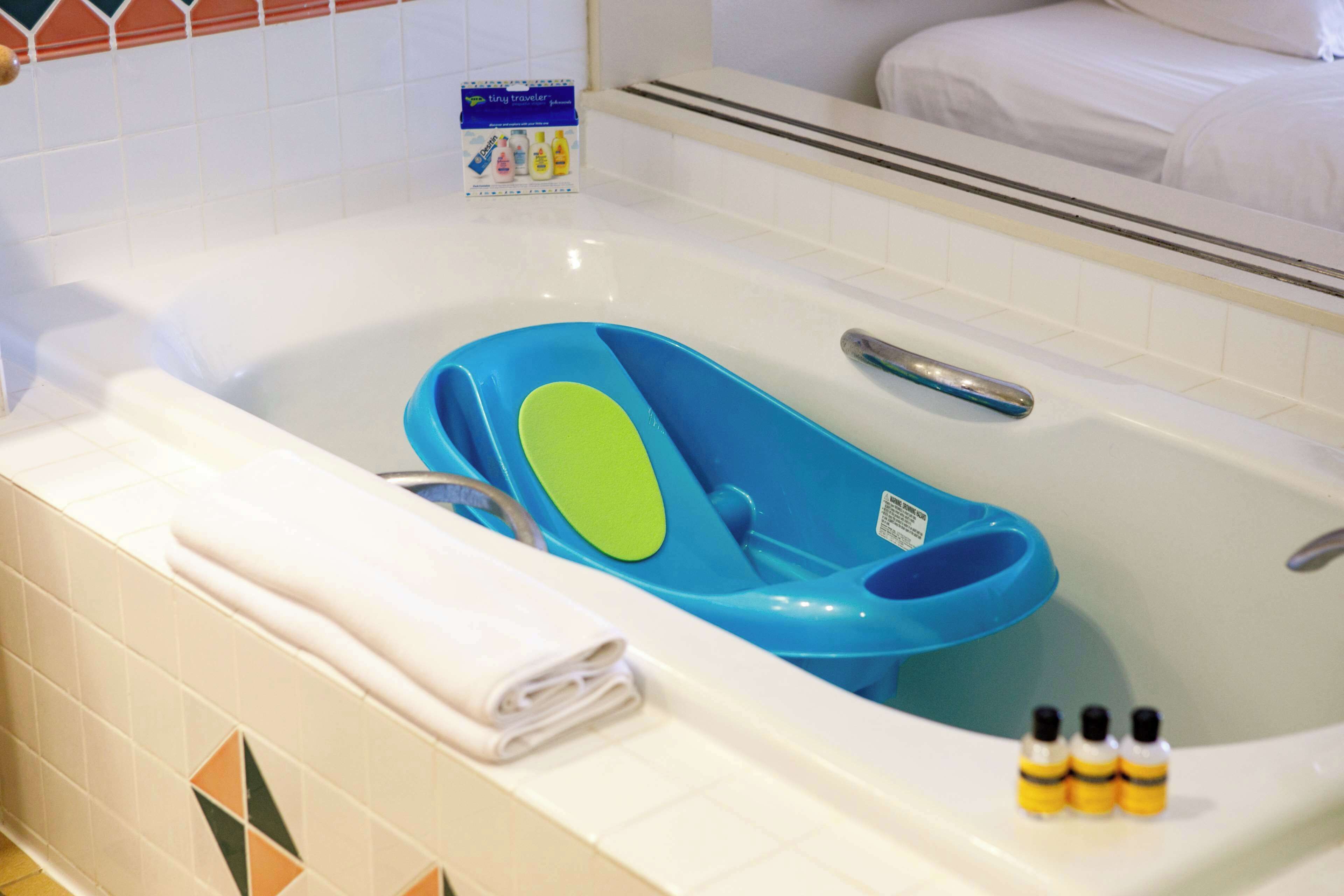 A bathtub with a blue and green seat, a rolled towel, and three small bottles.