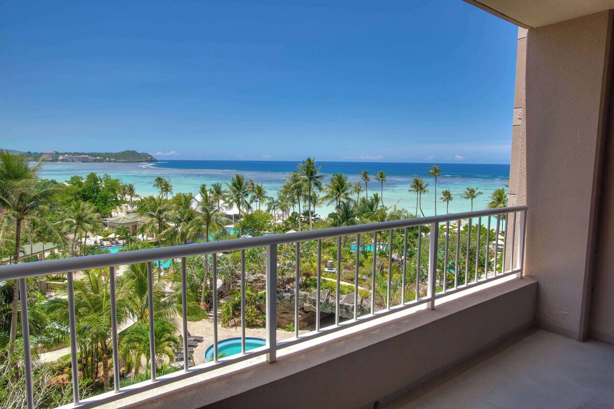 A balcony with a view of a beach, palm trees, and a swimming pool.