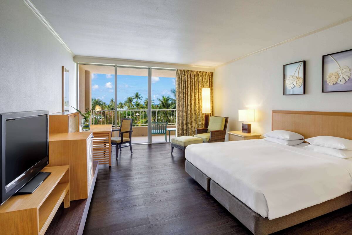 A hotel room with a bed, a TV, a desk with a chair, a balcony with a view of palm trees, and two framed artworks on the wall.