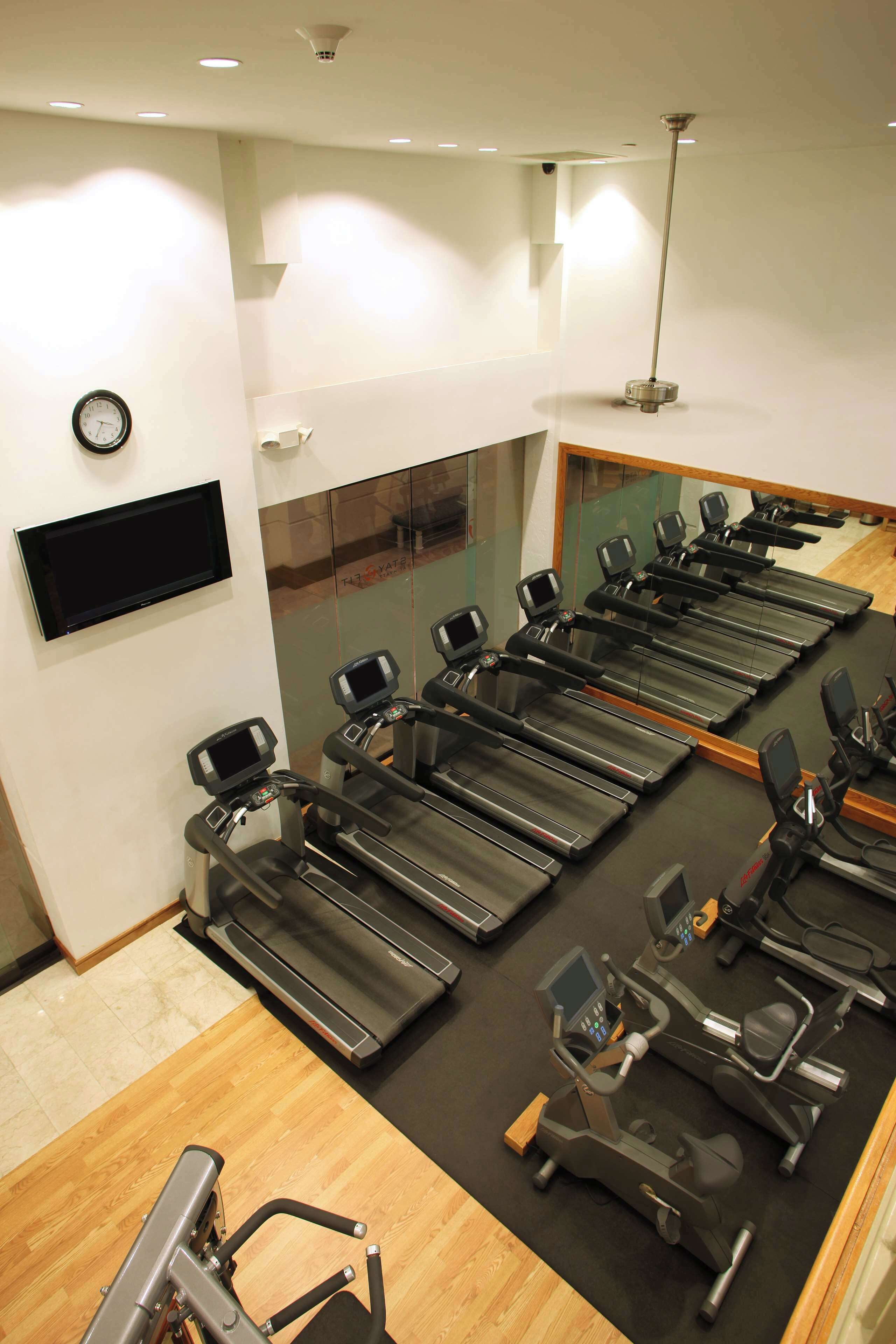 Une salle de sport équipée de plusieurs tapis de course et de vélos d’exercice, avec une horloge au mur et un grand miroir.