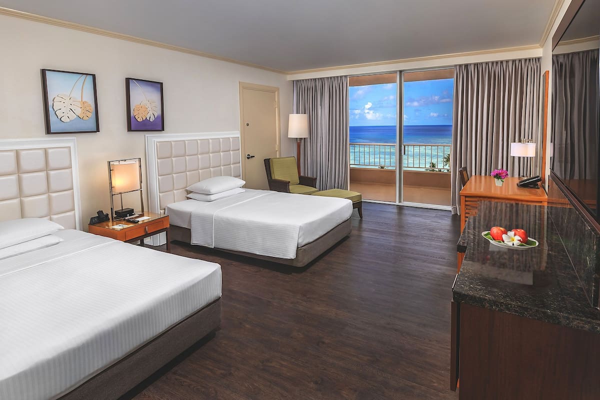 A hotel room with two beds, a desk, a chair, a nightstand, a lamp, and a view of the ocean.