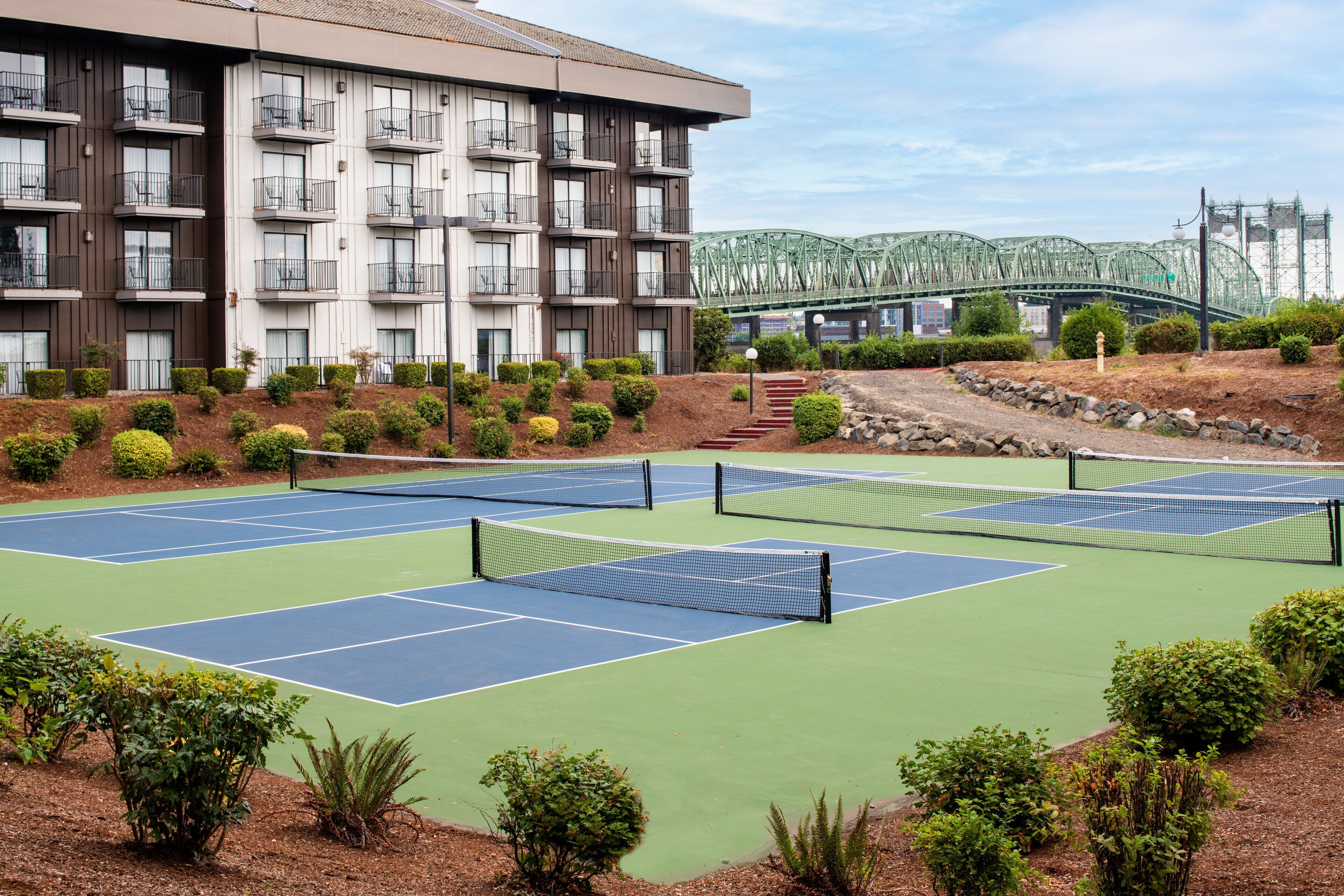 A tennis court complex with multiple courts, a building with balconies, and a bridge in the background.