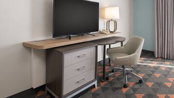 A hotel room with a flat-screen TV on a wooden desk, a chair, a lamp, and a telephone.