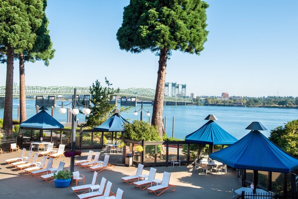 A waterfront patio with lounge chairs and umbrellas, overlooking a river and a bridge.
