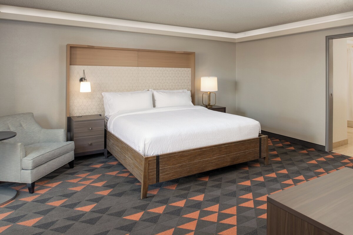 Une chambre d’hôtel avec un lit, une table de chevet, une lampe, une chaise et un tapis à motifs.