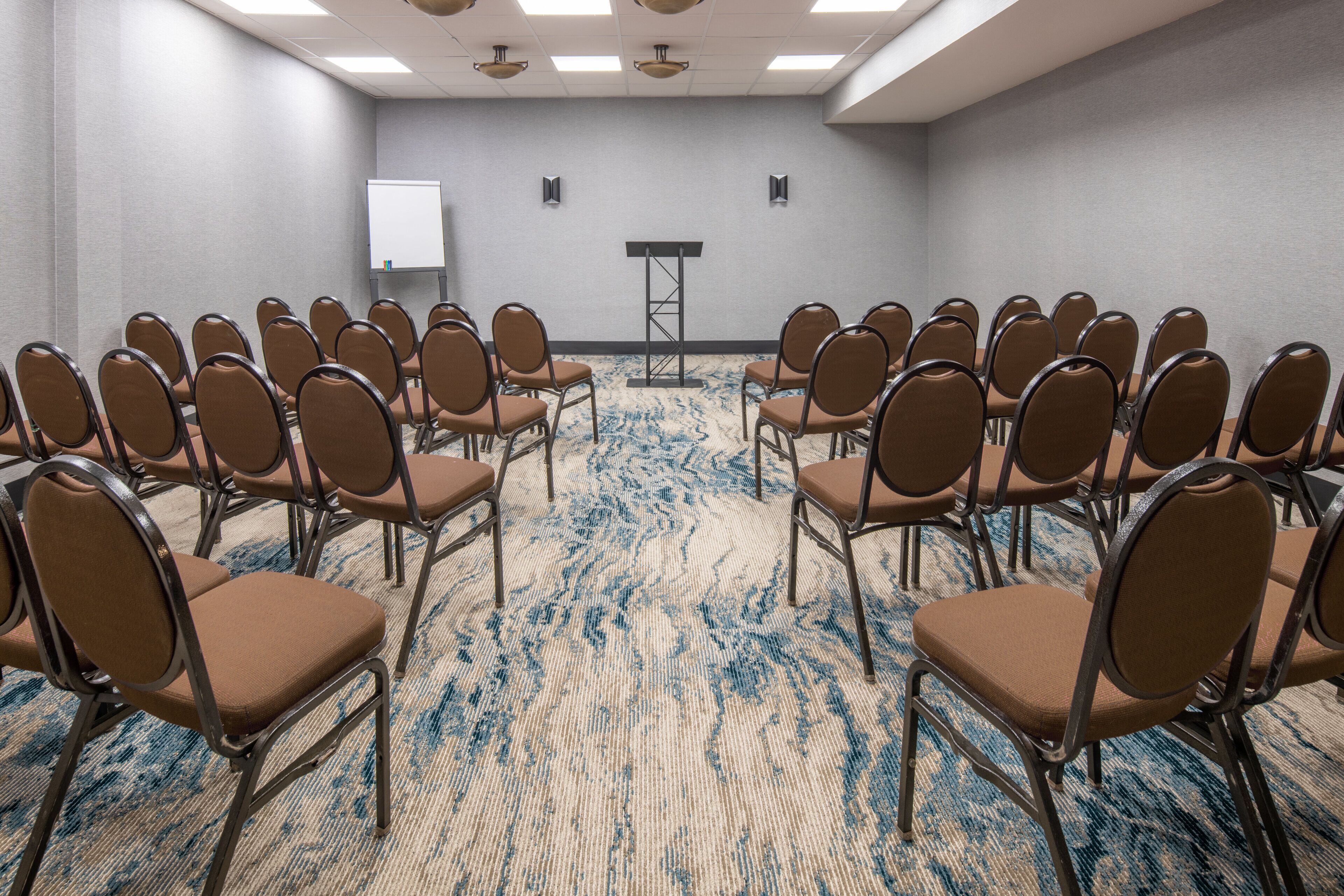 A conference room with rows of chairs facing a podium, a whiteboard, and two speakers on the walls.