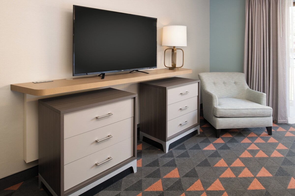Une chambre d’hôtel avec un téléviseur à écran plat posé sur une étagère en bois, une lampe, une chaise et un tapis à motifs.