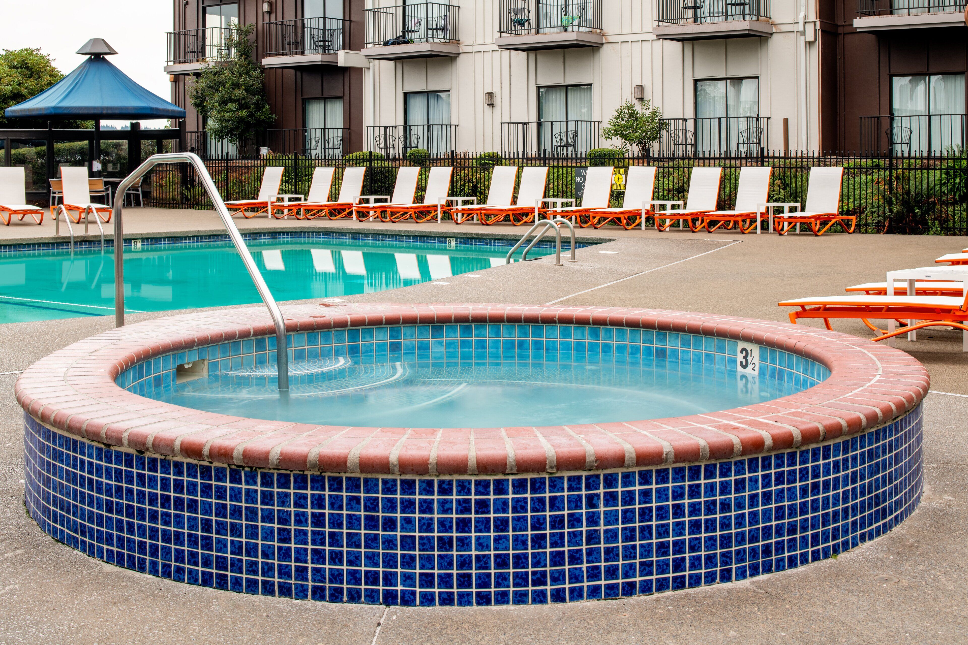 A hot tub with a tiled edge, surrounded by lounge chairs and a pool.