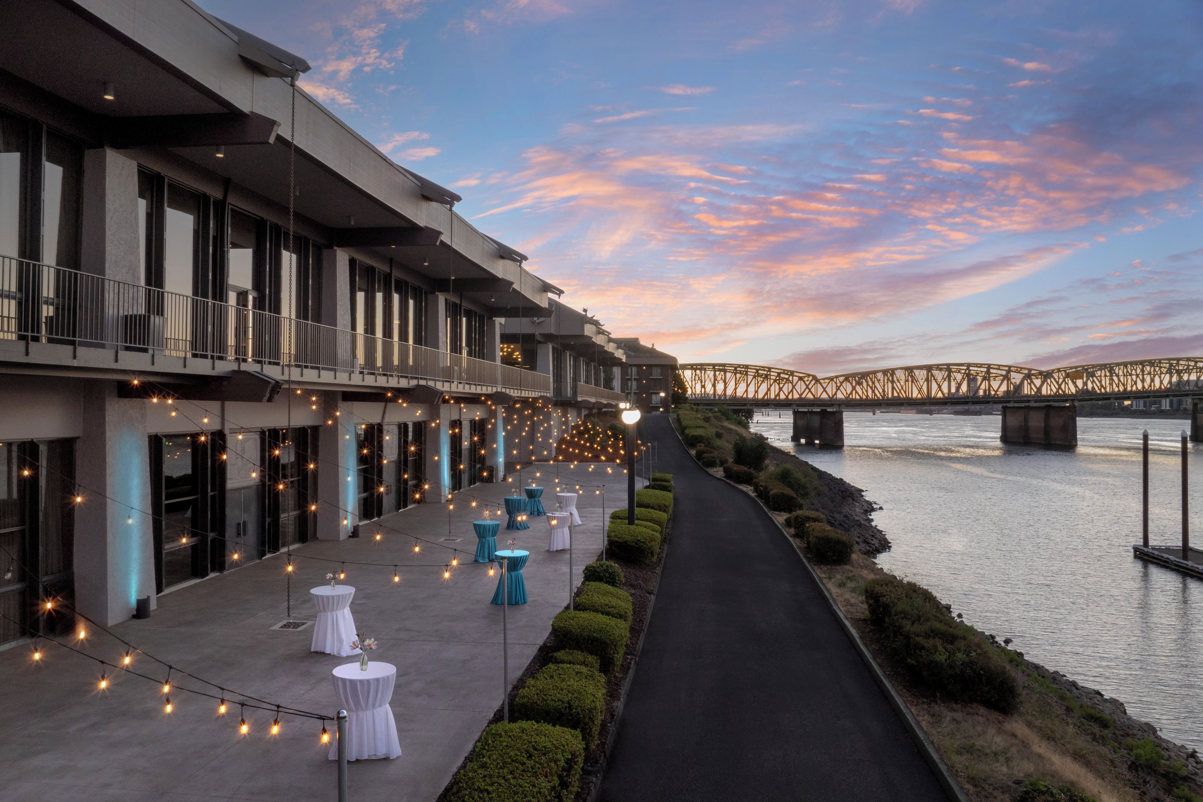 A riverside walkway with tables and chairs, a bridge in the background, and a modern building with balconies.