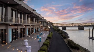 A riverside walkway with tables and chairs, a bridge in the background, and a modern building with balconies.