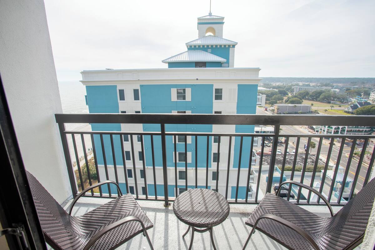 A balcony with wicker chairs and a small table, overlooking a blue and white building with a tower.