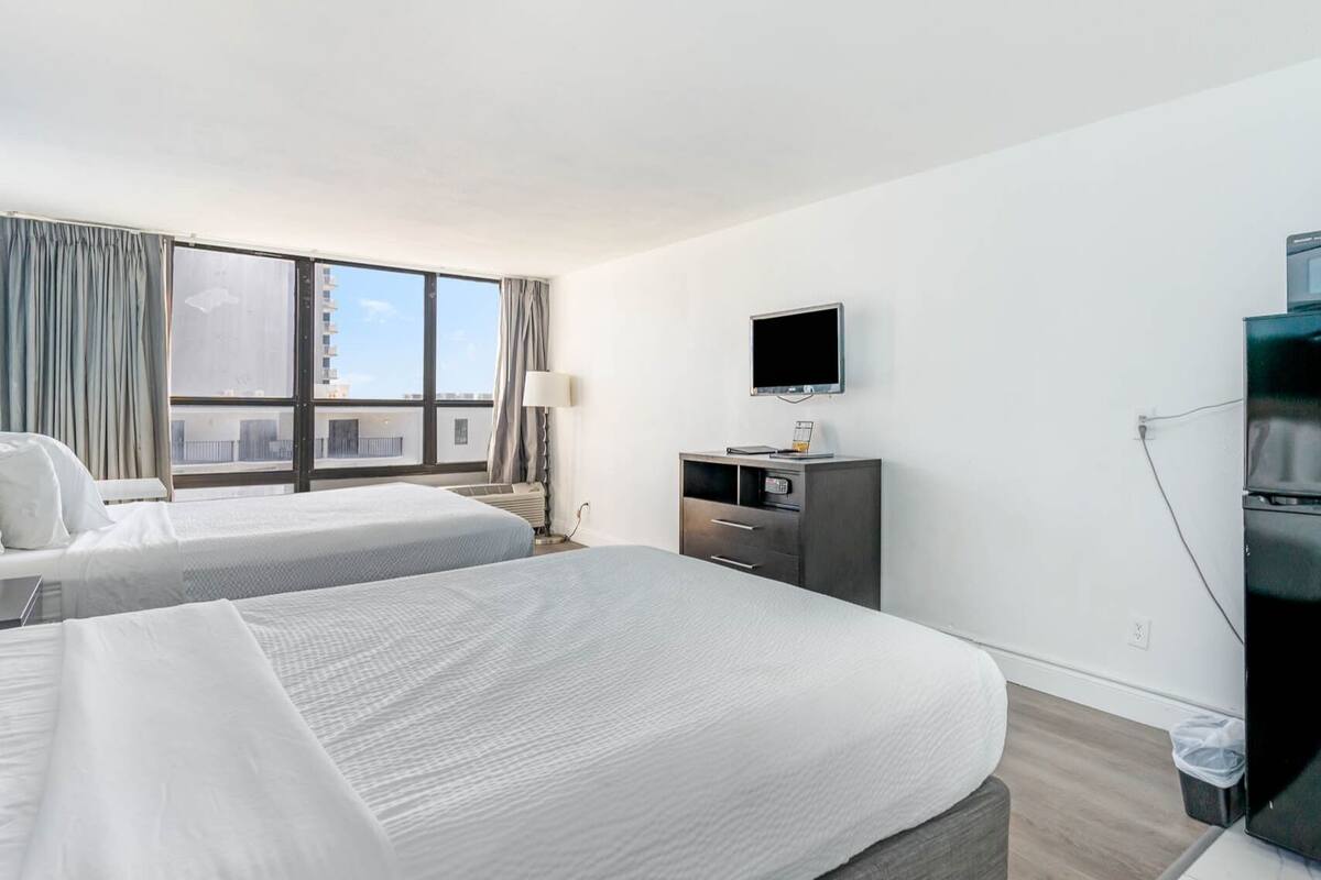A hotel room with two beds, a TV, a desk with a computer, and a window with curtains.