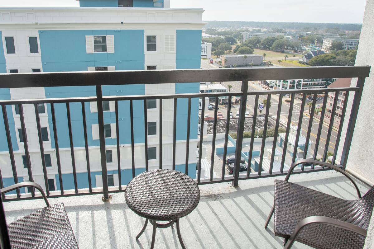 A balcony with a view of a building with a blue and white facade, a small table, and two wicker chairs.