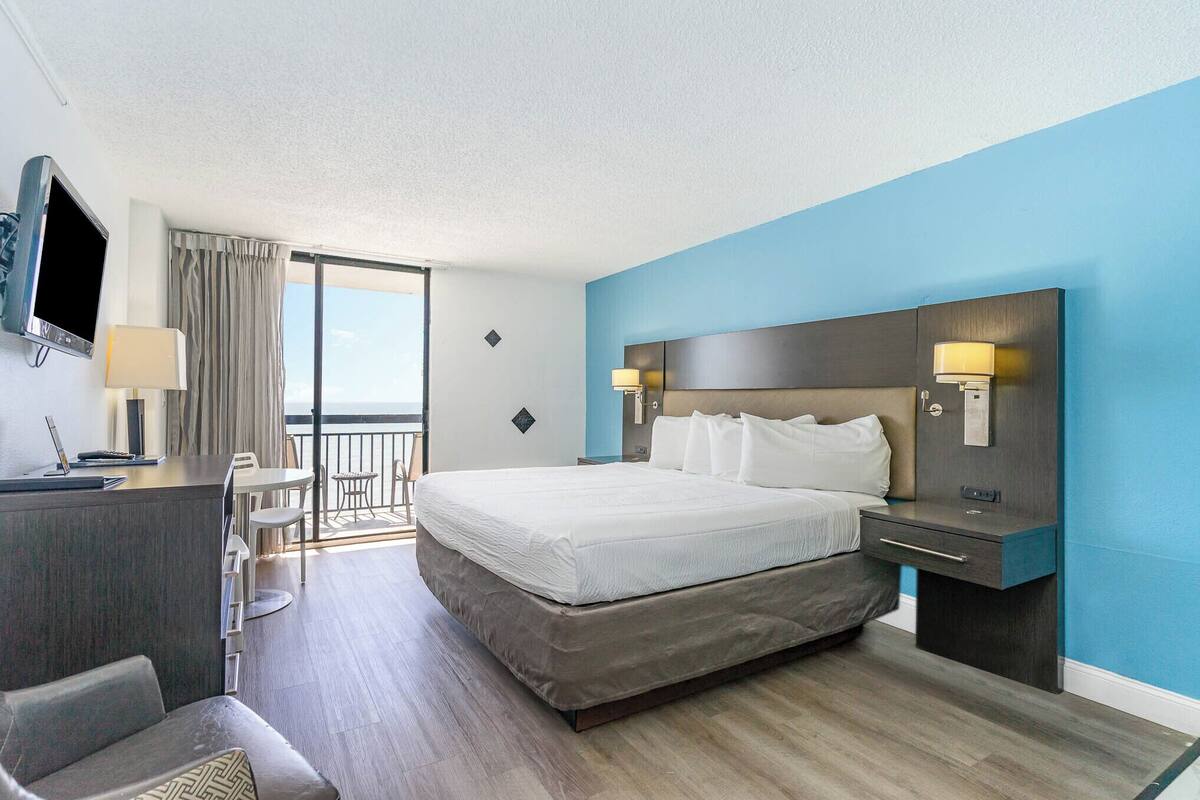 A hotel room with a bed, desk, TV, and a balcony with curtains.