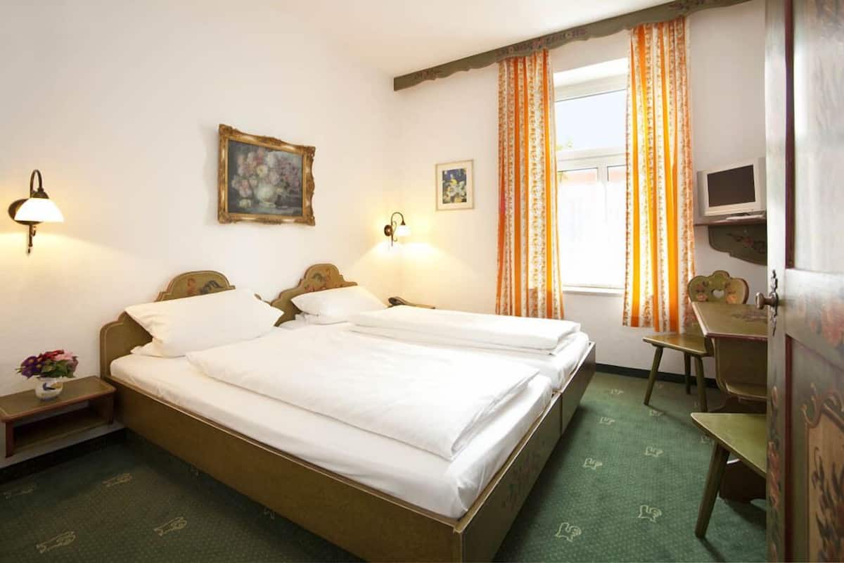 A hotel room with a double bed, two bedside lamps, a small table, a chair, a television, and a window with curtains.