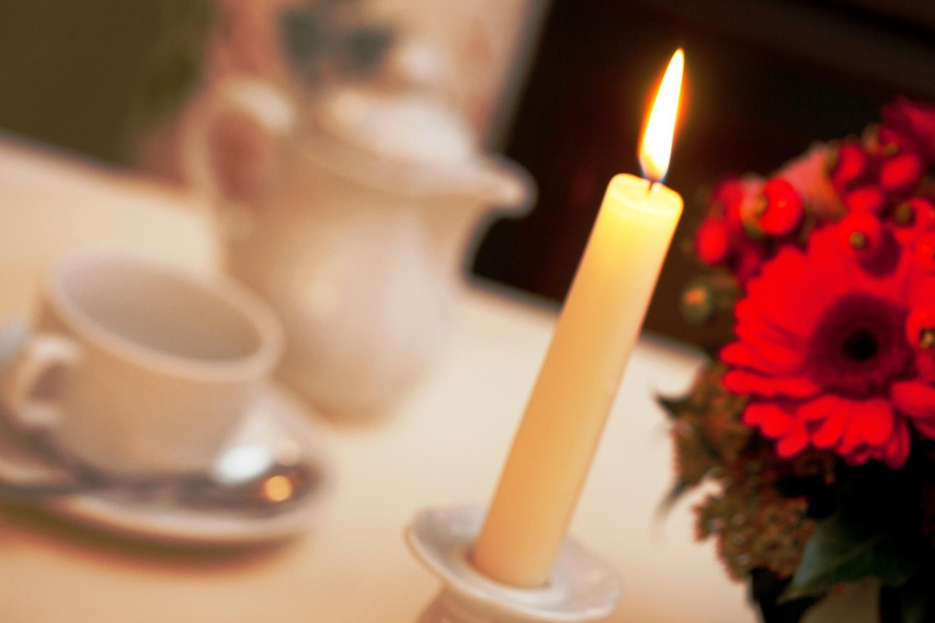 Primer plano de una vela encendida sobre una mesa, con una tetera y una taza al fondo.