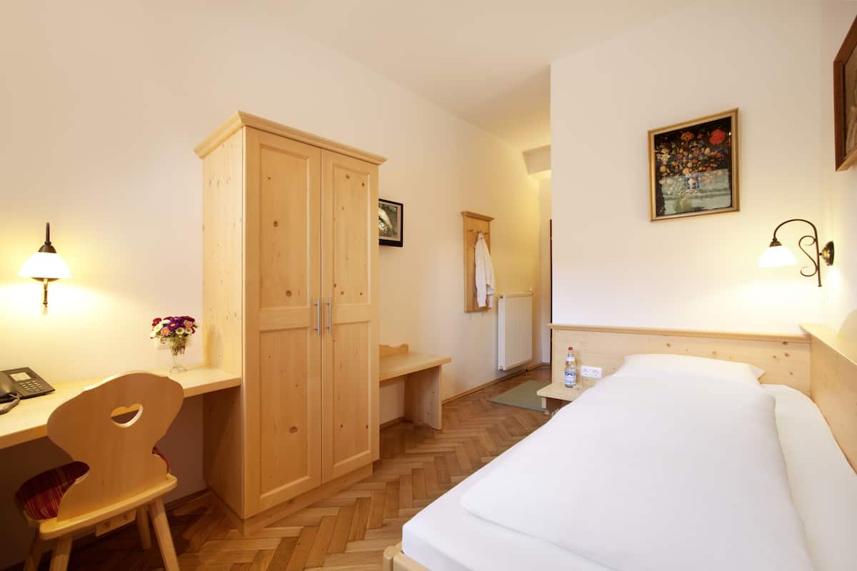 A hotel room with a bed, wooden wardrobe, desk, chair, and a painting on the wall.