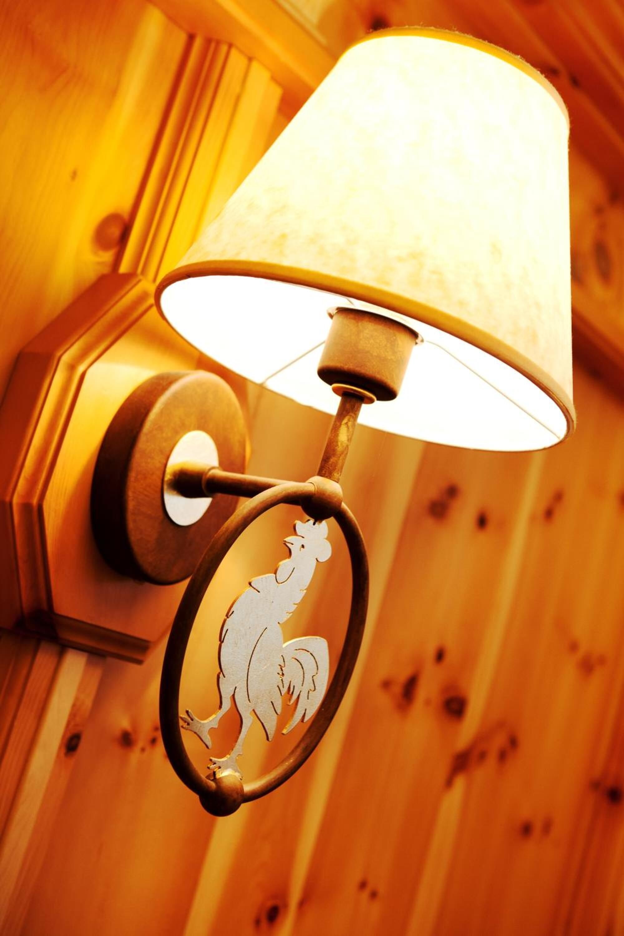 A wall-mounted lamp with a white lampshade and a decorative metal ring featuring a rooster design.