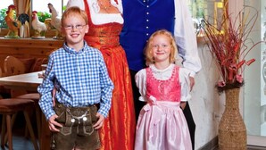 A family in traditional attire in a well-lit room with wooden beams, a dining table, and a vase with red branches. - Hahn Hotel (Munich)