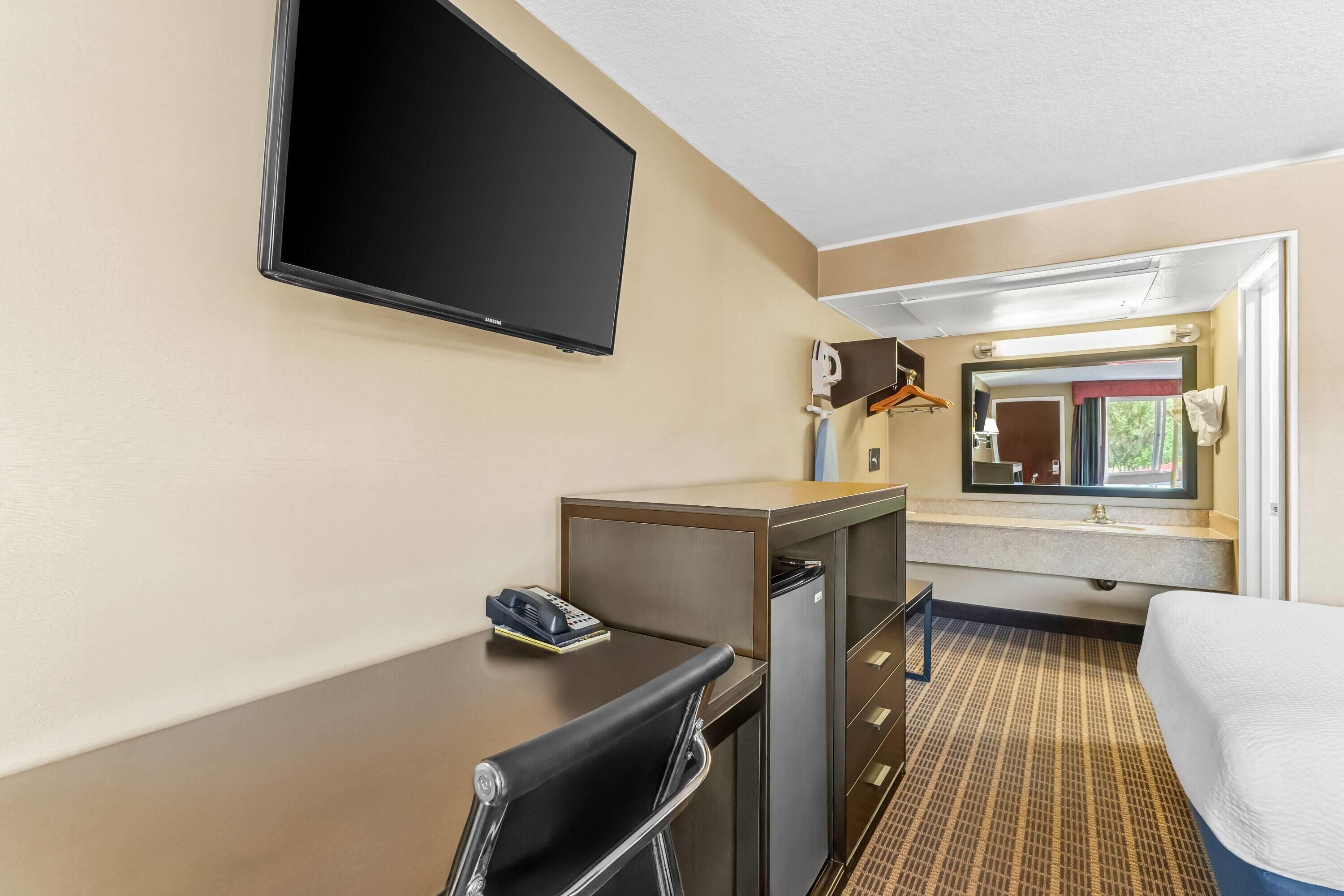 A hotel room with a bed, a desk with a chair, a television mounted on the wall, a small table with a phone, and a window with a view of the outside.